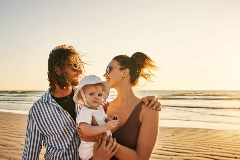 Young family on the beach with parents looking happy with baby in their hands knowing they have overcome first time parents struggles