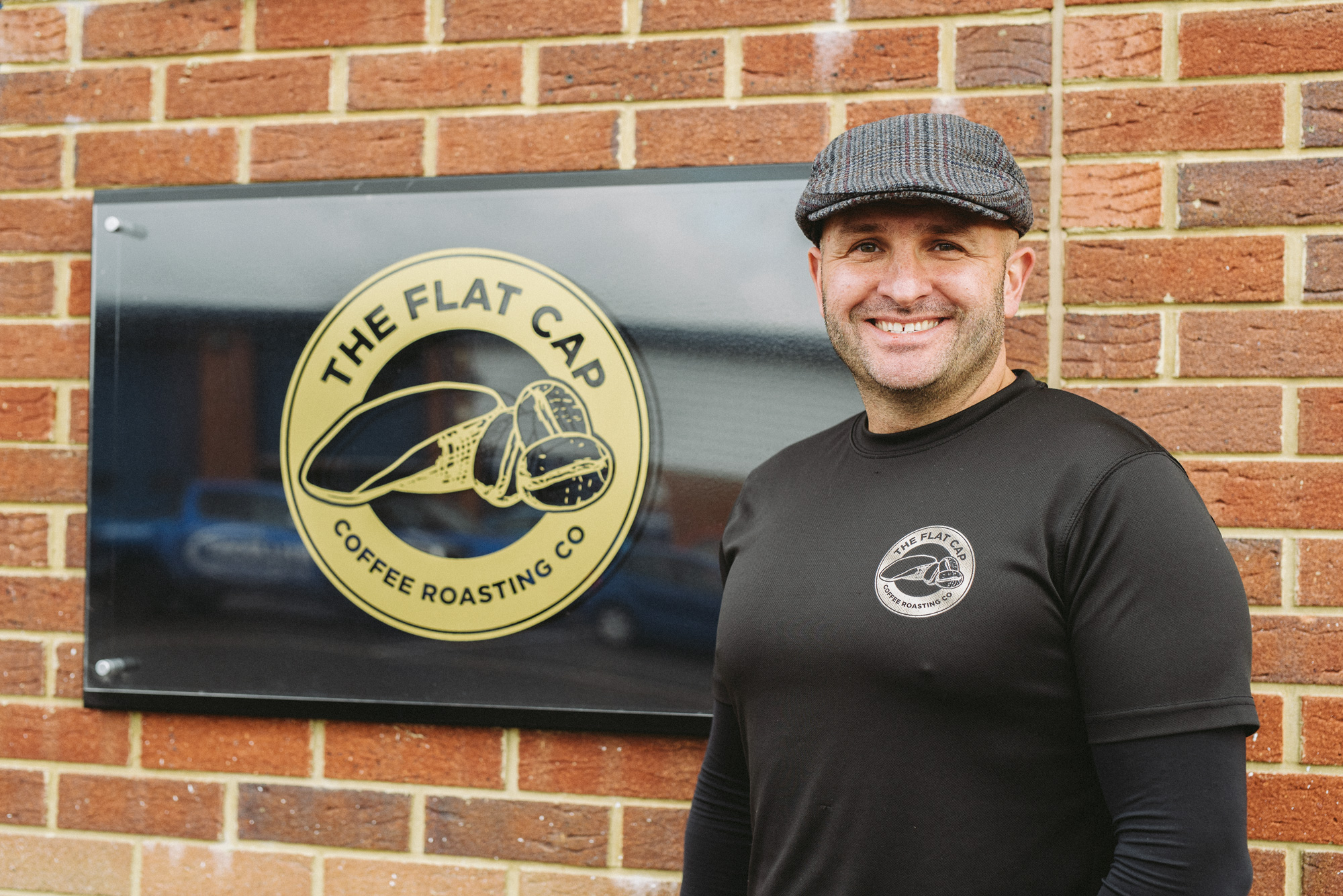Mark, founder of Flat Cap Coffee Roasting Co. stood next to a Flat Cap Coffee sign on a wall