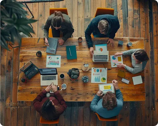 Aerial view of five people sitting at a table with laptops and reports spread around the table.
