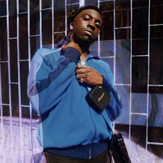 Kelvin Krash in a blue jacket leaning against a tiled wall with a Marshall Willen portable speaker.