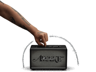 A tattooed hand adjusts the volume on a Marshall Acton III Awake NY Edition speaker, nestled against a black background.