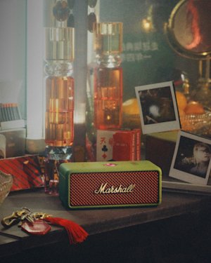 An Emberton III Lunar New Year 2025 Edition portable speaker by Marshall sits on a cluttered desk, surrounded by an eclectic mix of photographs, keychains, cards, and colorful bottles.