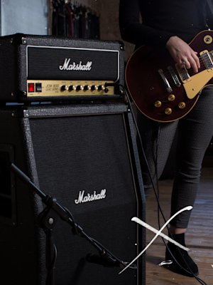 A woman is playing a guitar in front of a Marshall SC20H Head.