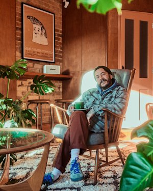 Un hombre con barba sentado en un sillón, con una taza en la mano, en una acogedora habitación llena de plantas, con una decoración elegante y obras de arte enmarcadas en la pared.