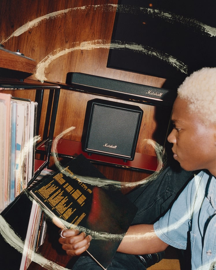 A person with short blonde hair examines a vinyl record sleeve near shelves of records, and a Marshall Heston Sub 200 Black subwoofer.