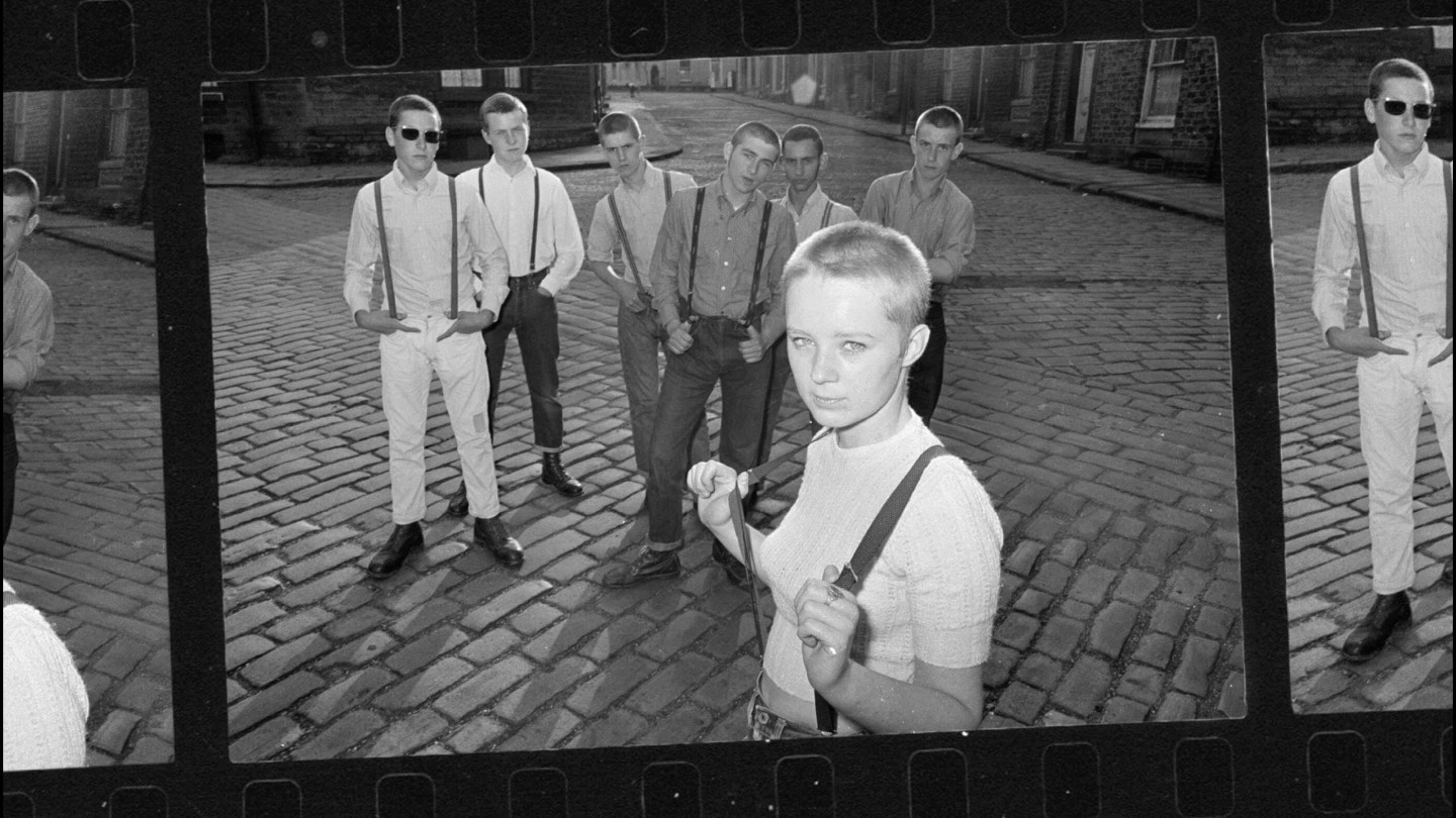 Mujer joven en primer plano con un grupo de hombres jóvenes detrás de ella en una calle adoquinada, todos vestidos a la moda de los años sesenta.