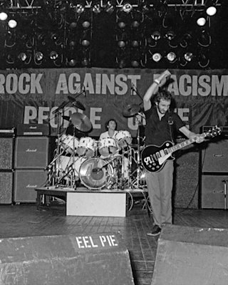Black and white photo of a guitarist and drummer performing on stage at a rock against racism concert, with banners and stage equipment visible.