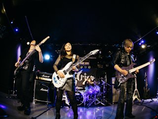 A four-member rock band performs on stage, with two guitarists, a bassist, and a drummer. The stage is lit with blue and purple lights, and there is a banner with the band's name in the background.