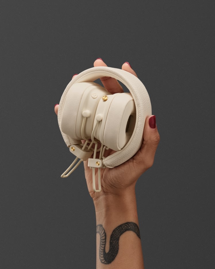 Eine Person mit rotem Nagellack und einem Tattoo am Handgelenk hält einen Marshall Major V Cream On-Ear-Kopfhörer vor einem grauen Hintergrund.