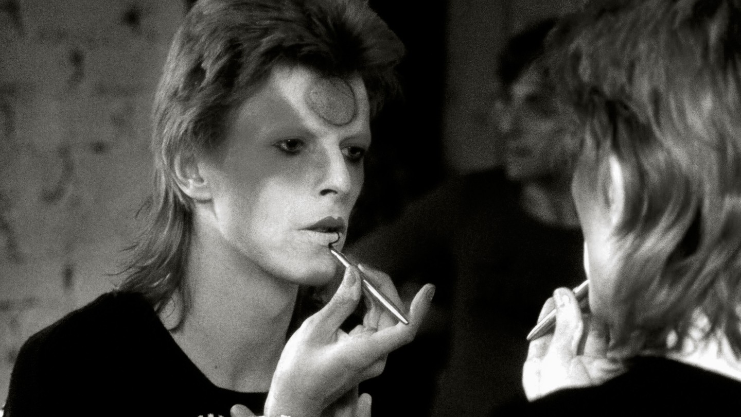Photo en noir et blanc d'une personne au maquillage et à la coiffure spectaculaires, allumant une cigarette tout en regardant au loin d'un air pensif.