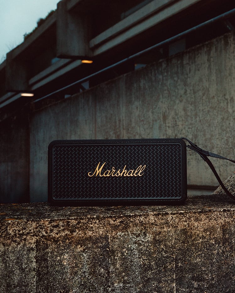 Marshall Middleton II Black and Brass portable Bluetooth speaker sits on a concrete ledge with a concrete structure and lights in the background.