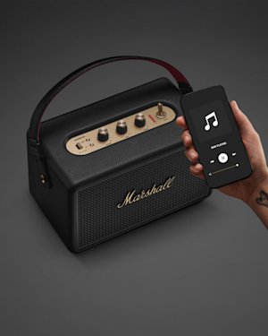 Una mano sostiene un teléfono delante de un altavoz Marshall Kilburn III negro y latón.