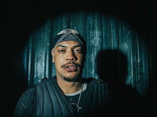 A man wearing a dark bandana and a tactical vest stands in a dimly lit environment with a visible spot of light.