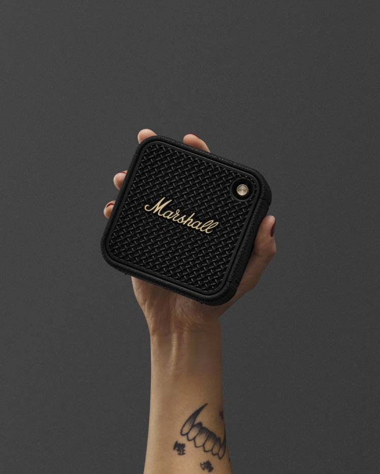 A person holds Marshall Willen II Black and Brass portable bluetooth speaker against a dark background.
