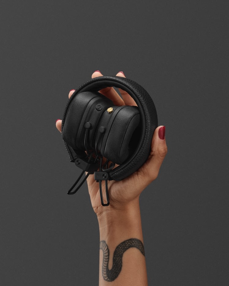 Woman holding a pair of folded black Marshall Major V on-ear headphones against a grey background.