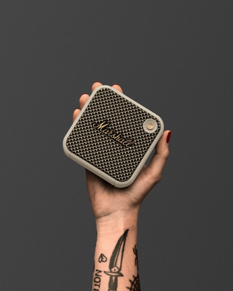 Une main tatouée tenant une enceinte portable Marshall Willen Cream. Facile à transporter, le Marshall Willen est un petit haut-parleur pratique.