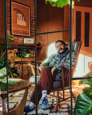 Un hombre se sienta en un sillón con una taza en la mano, rodeado de plantas en una habitación acogedora y cálidamente iluminada con un cuadro enmarcado en la pared.