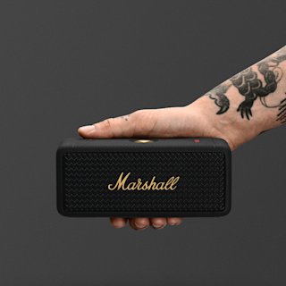 Une enceinte Bluetooth portable Marshall Emberton II en noir et laiton, avec un bouton de commande et un logo en laiton, posée sur une surface sombre et unie.