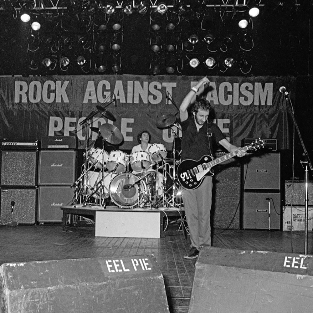 Una banda actúa en el escenario de un concierto de "rock contra el racismo"; el batería al fondo, el guitarrista tocando enérgicamente en primer plano.