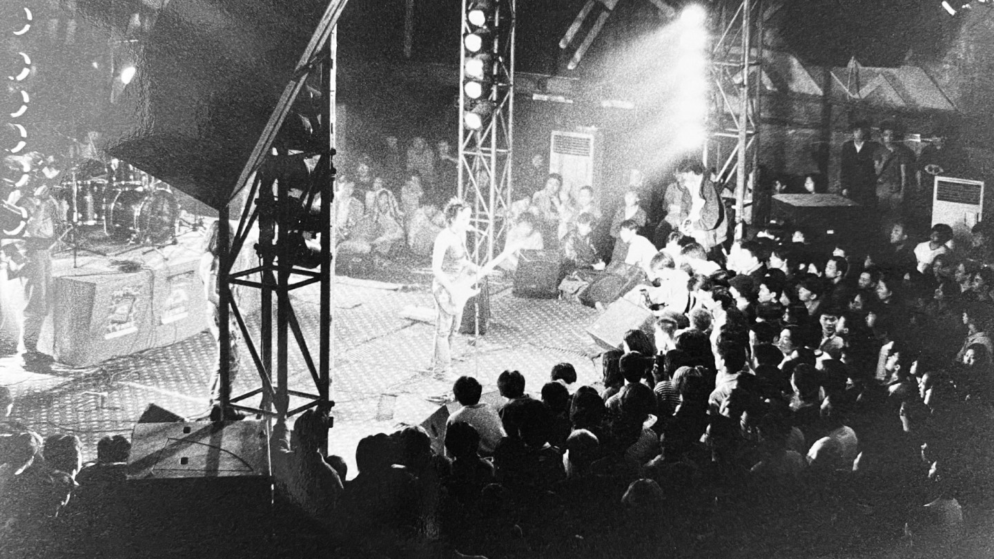 明るい照明のステージでギターを弾くミュージシャンと、薄暗い会場でそれを見守る観客。