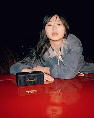 Woman lying on a red surface next to a Marshall Emberton II outdoor speaker at night.