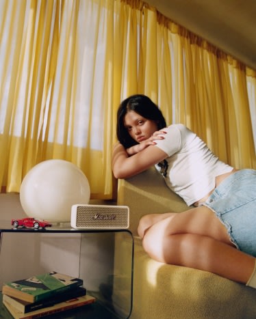 Suki leans on a chair next to a table with Emberton III Cream portable speaker from Marshall.