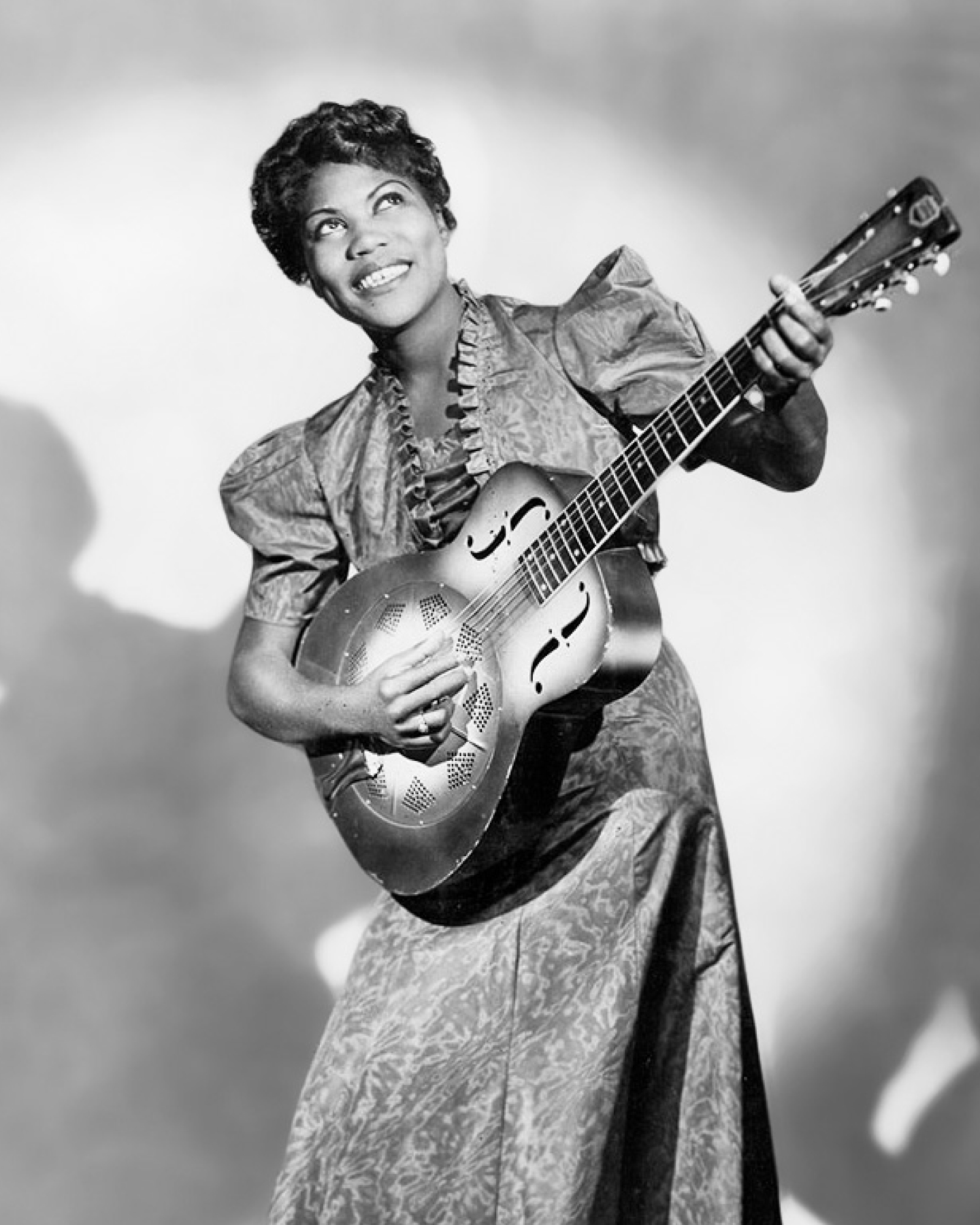 Photo en noir et blanc d'une femme souriante tenant une guitare à résonateur, vêtue d'un chemisier à volants et d'une robe à motifs.