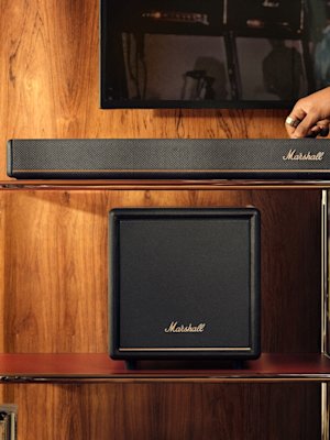 A hand adjusts a Marshall soundbar on a wooden shelf, with the Marshall Heston Sub 200 subwoofer placed below.
