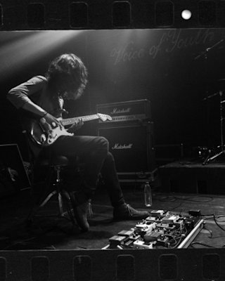 Imagen en blanco y negro de un guitarrista sentado en un taburete con una pedalera a sus pies.
