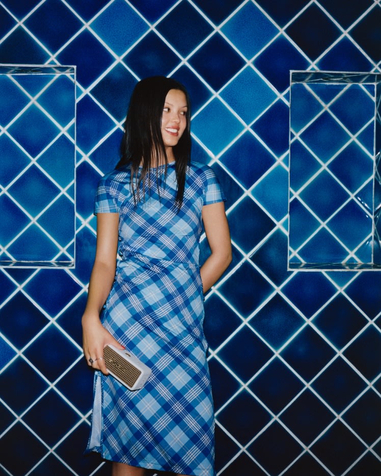 A woman in a blue plaid dress holding an Emberton III Cream portable bluetooth speaker by Marshall.