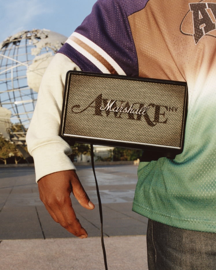 A person holding a Marshall Acton III Awake NY Edition home speaker under their arm stands near a globe sculpture, wearing a multicolored shirt.