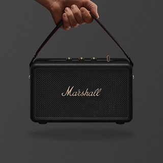 Una mano sujeta por el asa un altavoz portátil Marshall Kilburn III en negro y latón sobre un fondo oscuro liso.