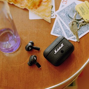 A pair of black wireless earphones and their charging case are on a wooden table, alongside playing cards, keys, and a glass.