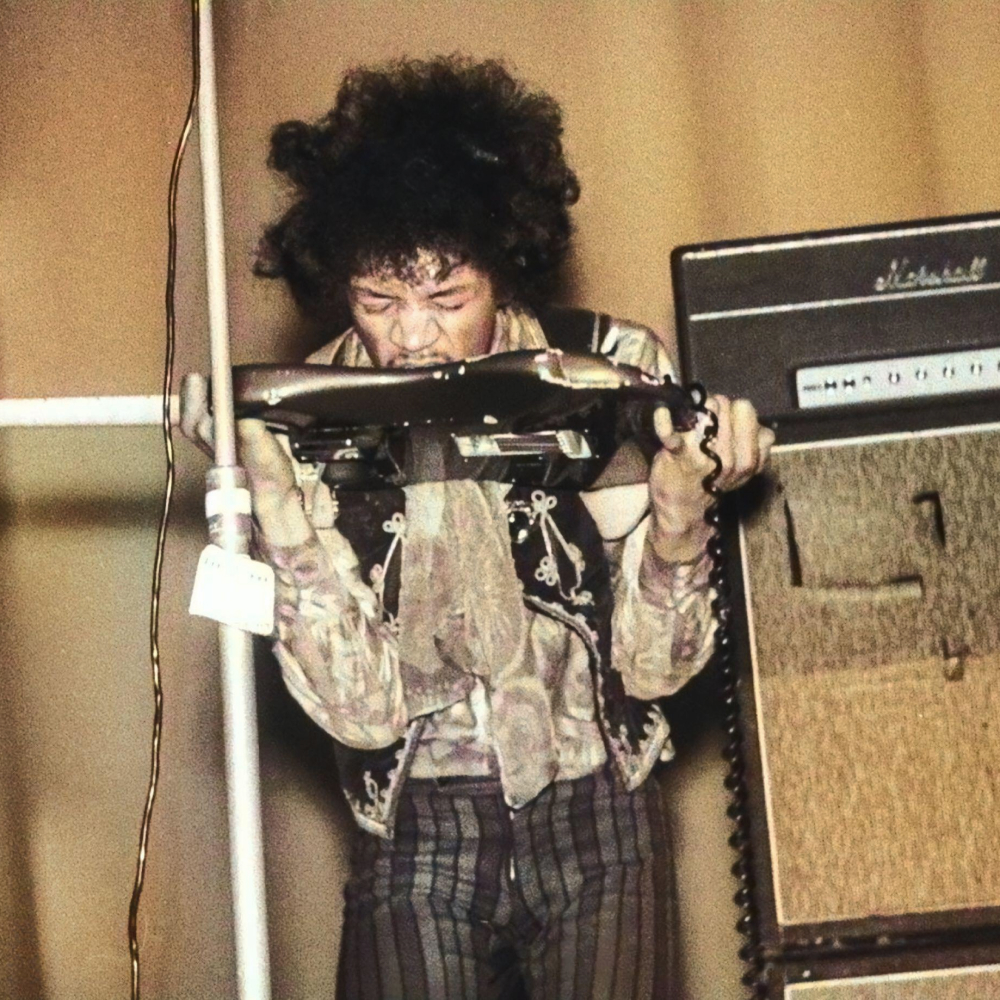 Image en couleur de Jimi Hendrix debout devant un amplificateur Marshall. Il porte la guitare à sa bouche et en joue avec ses dents.