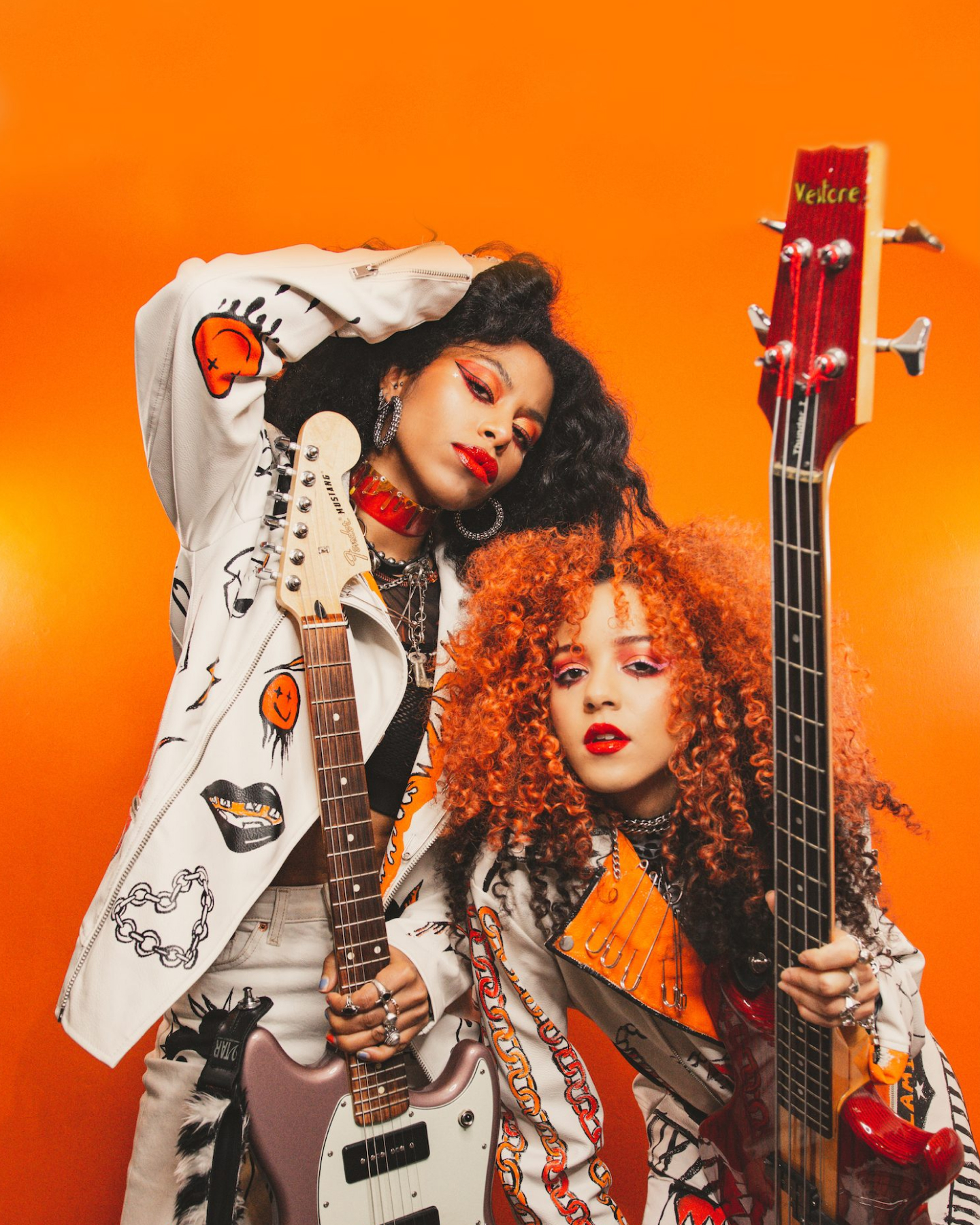 Zwei stilvolle Musiker, einer in einer weißen Jacke und der andere in einer orangefarbenen Bluse, posieren mit E-Gitarren vor einem orangefarbenen Hintergrund.