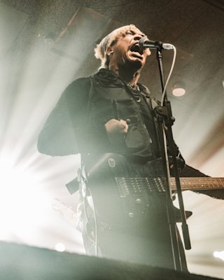 A musician passionately sings into a microphone while playing an electric guitar on stage. Bright lights shine from behind, creating a dramatic effect.