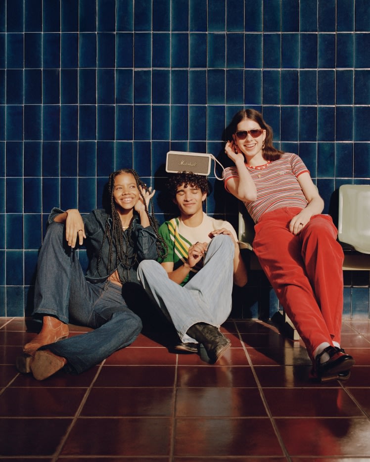 A group of people sitting on the floor in front of a blue tiled wall, with a white Marshall Middleton Cream speaker placed prominently amidst them.