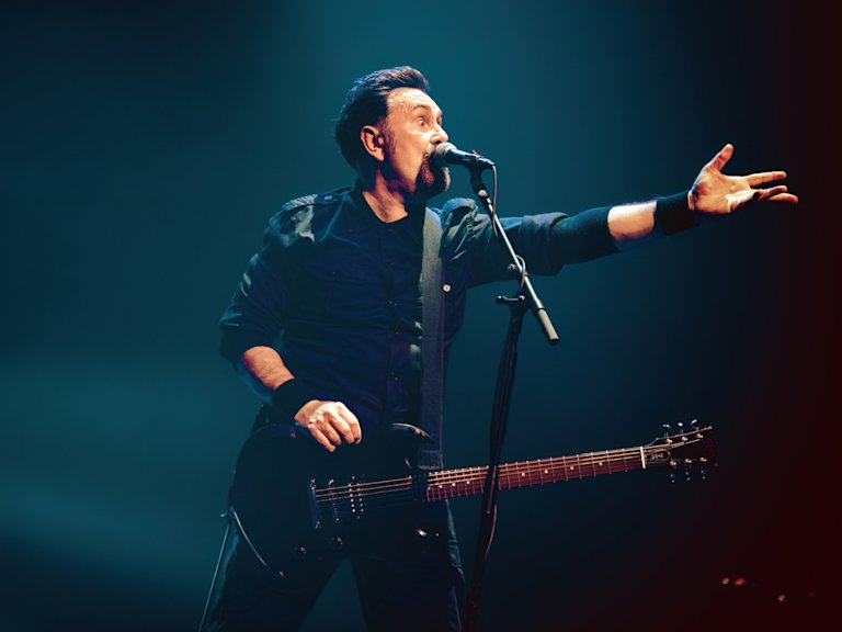 A musician performs on stage with a guitar, holding a microphone and gesturing with one hand.