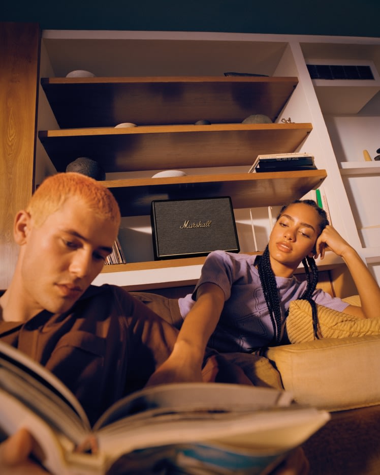 Ein Mann und eine Frau genießen die außergewöhnliche Klangqualität des Marshall Woburn III Black-Lautsprechers, während sie gemeinsam ein Buch auf ihrer gemütlichen Couch lesen.
