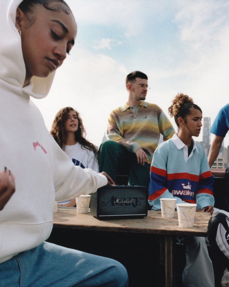 A group of people gather around a wooden table with a Marshall Acton III Awake NY Edition bluetooth speaker and paper cups on it.