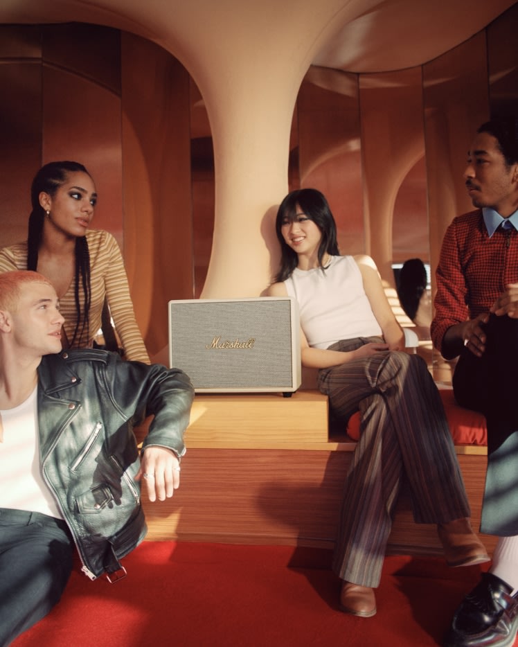 A group of people sitting on a couch listening to music on a Marshall WOBURN III CREAM speaker.