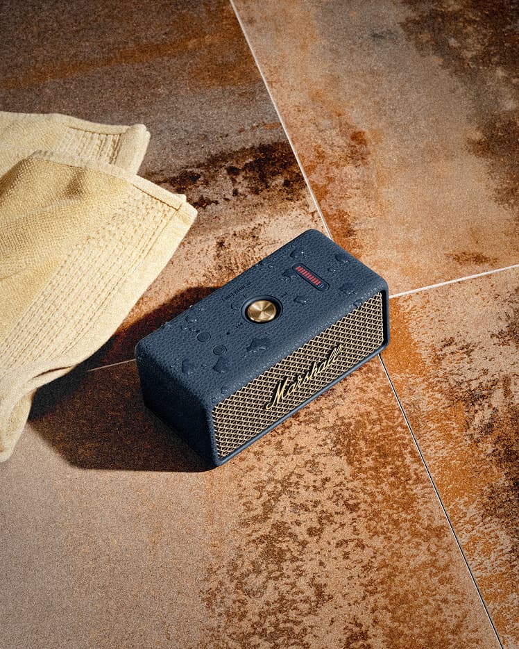 Marshall Emberton III Midnight Blue portable Bluetooth speaker sits on brown tiles next to a yellow towel, with water droplets visible on its surface.