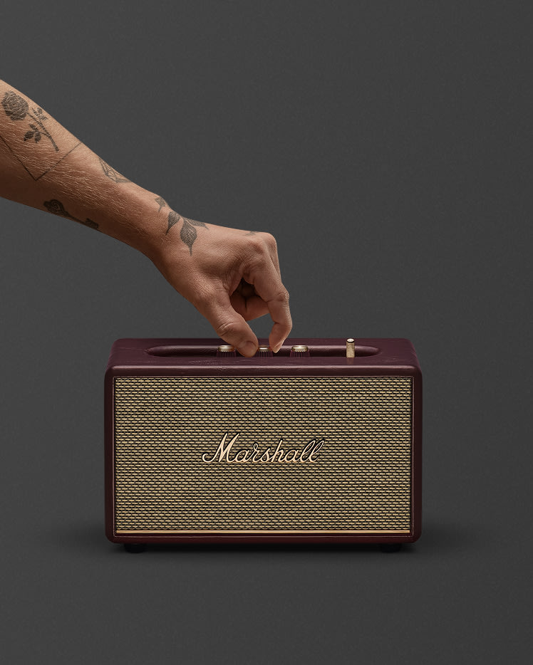 A person with tattooed hands adjusts a knob on a Marshall Acton III Burgundy speaker against a plain gray background.