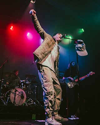 Musician Jarki Monno performs energetically on stage with a microphone, wearing a casual outfit and cap, amid colorful lights and a backing band with a drummer and bassist.