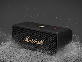 Marshall Emberton III Black and Brass speaker on a textured surface with small particles scattered around.