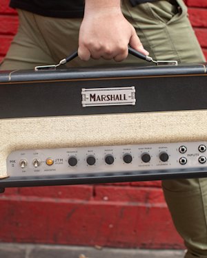 Una persona camina delante de un muro de ladrillos rojos portando el amplificador de cabezal Studio JTM