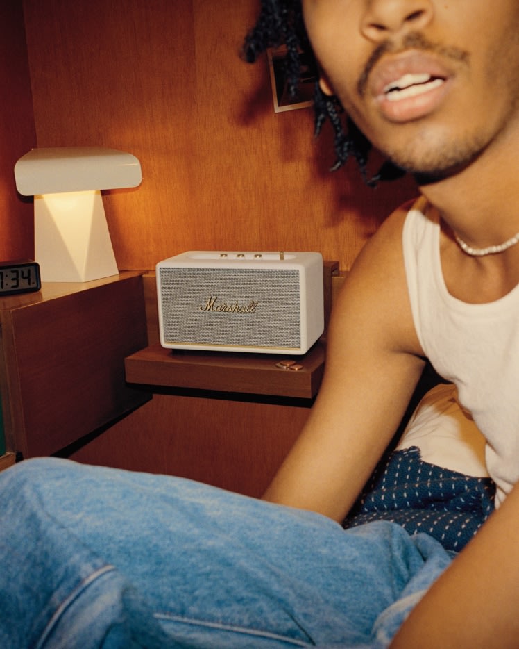 Ein Mann mit Dreadlocks sitzt auf einem Bett mit einem Marshall ACTON III CREAM Lautsprecher.