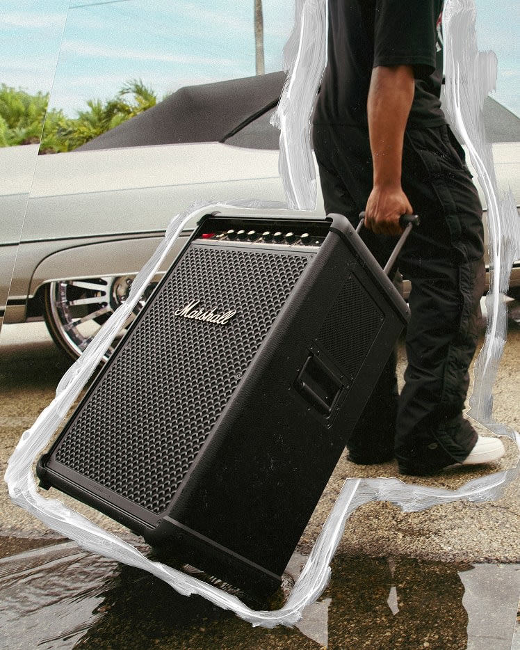 A person pulls a Marshall Bromley 750 party speaker next to a silver car parked outside.