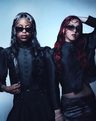 Dos estilosas artistas de Nova Twins posando delante de un árbol lleno de flores rosas.