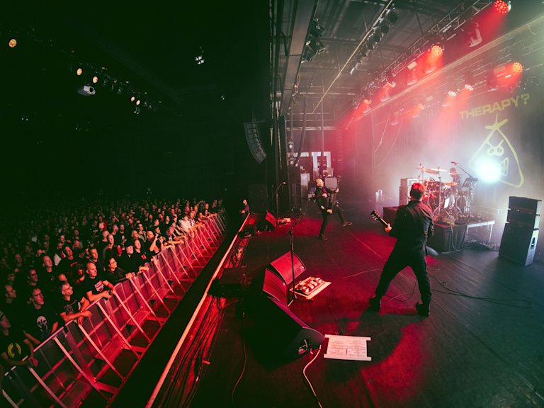 A band performs live on stage with guitars and drums under red lights infront of a large crowd. The backdrop displays the text "THERAPY?.